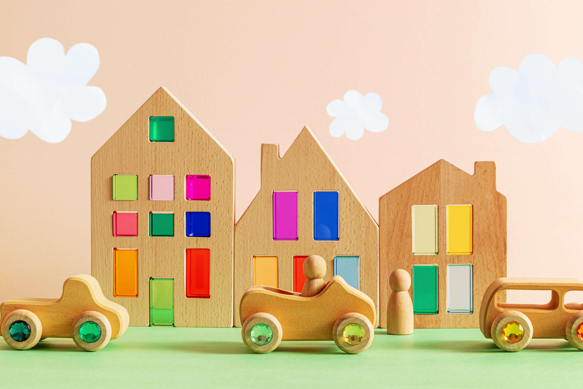 Otto wooden toy vehicles with gemstone wheels in front of colorful wooden houses and playful clouds.