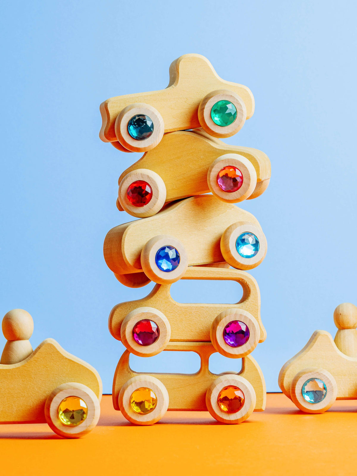 Otto wooden toy vehicles with gemstone wheels stacked playfully in front of a blue backdrop.