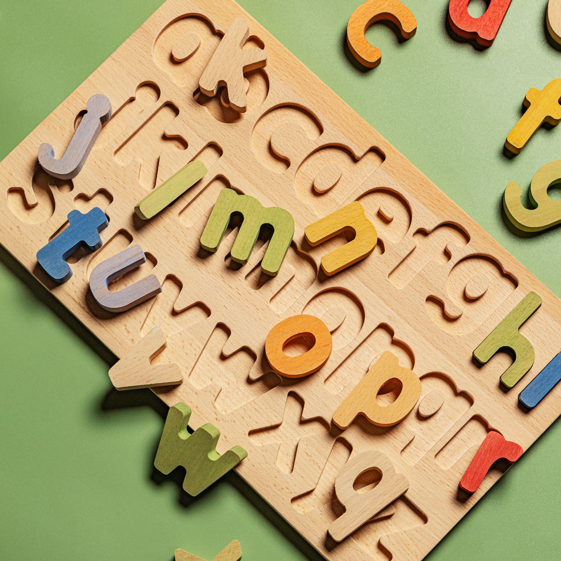 A wooden alphabet puzzle board with colorful alphabet letter pieces scattered on a solid green background.