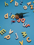 A small dark green dinosaur toy lies on a bright blue background among scattered colorful alphabet letters spelling out words like dinosaur.