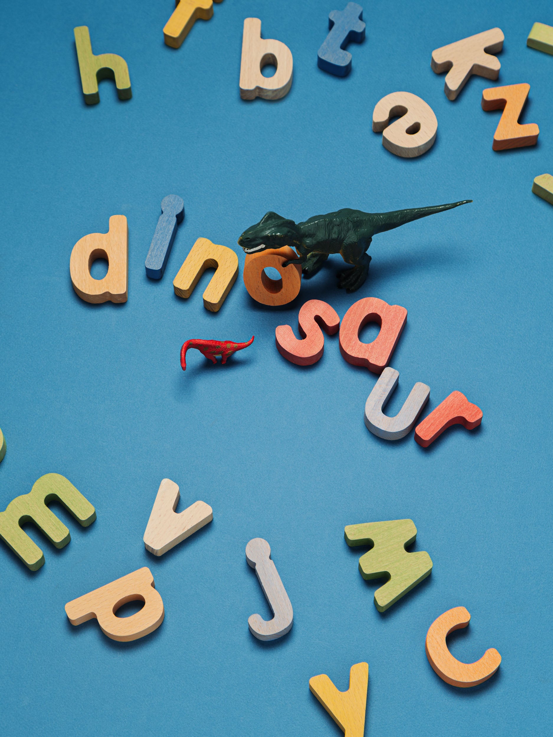 A small dark green dinosaur toy lies on a bright blue background among scattered colorful alphabet letters spelling out words like dinosaur.