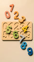 A wooden number puzzle board with carved slots and colorful loose number pieces on a warm beige background.